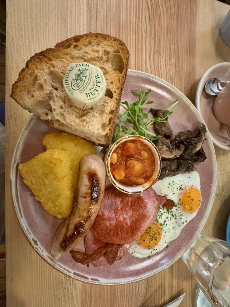 Full English breakfast featuring sourdough toast, homemade beans, fried eggs, hashbrown triangles, sausages, and bacon rashers served on a pottery plate.