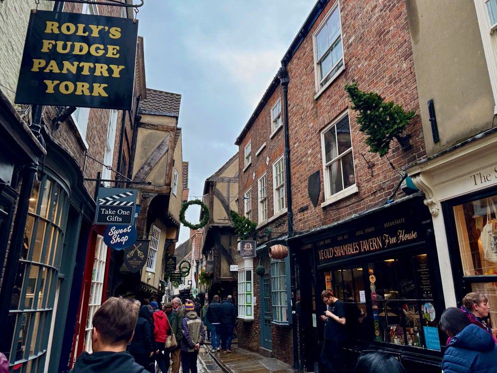 Half timber buildings and street signs hang over a narrow cobblestone street filled with people in central York