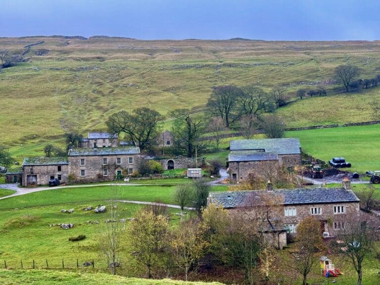 A Victorian farm with multiple stone buildings nad farm equipment in the yard tucked into a Yorkshire Dales hillside
