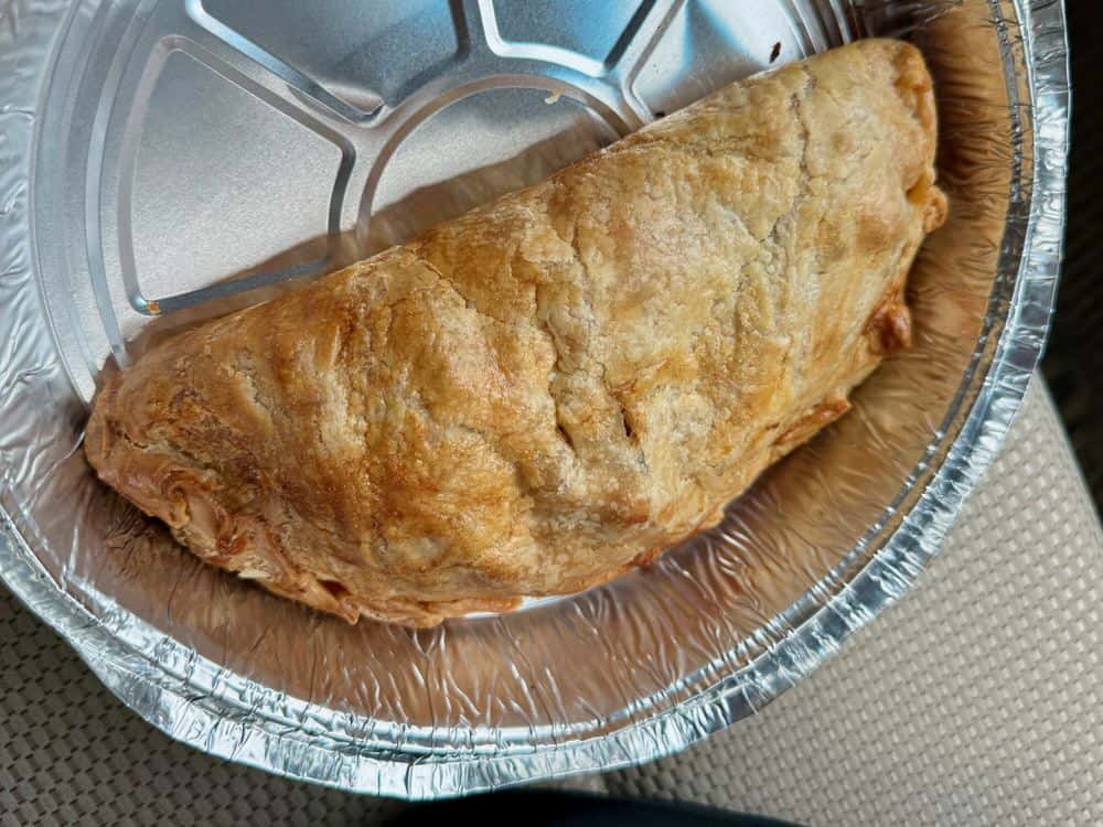 A baked Cornish pasty sits on an aluminum tin.