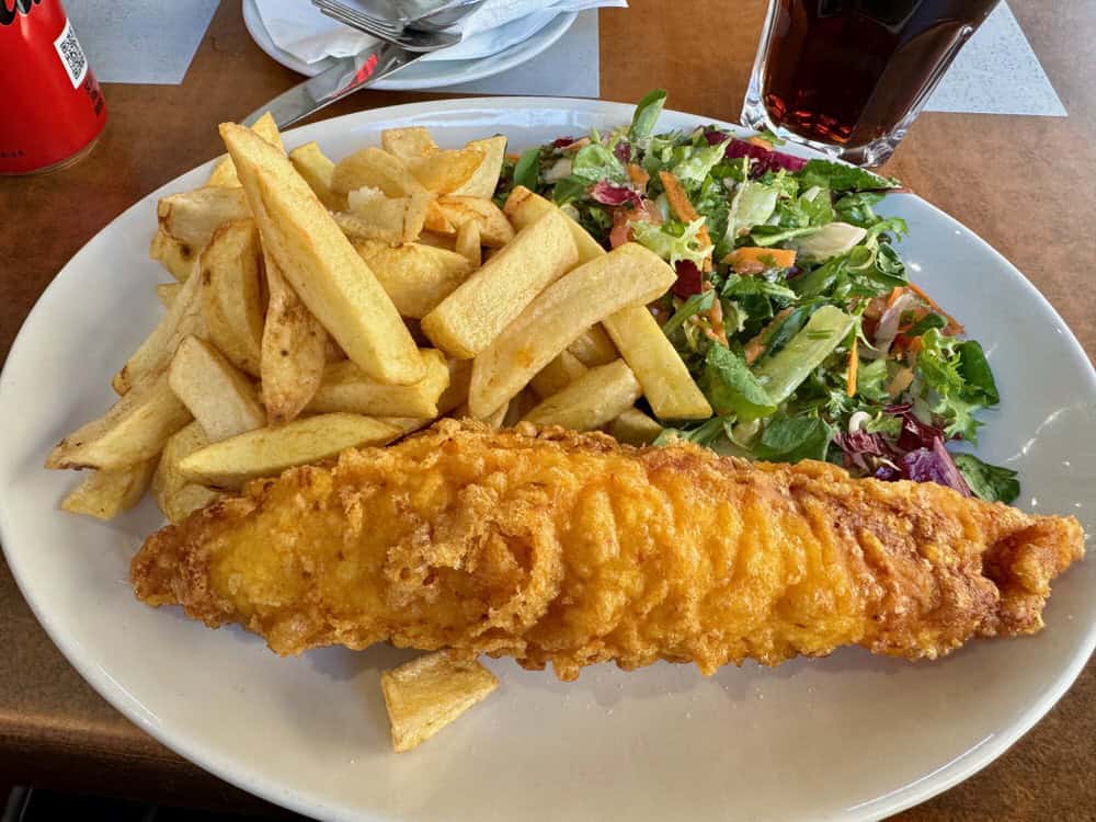 White plate covered with a large piece of fried fish, french fried potatoes, and a mixed salad