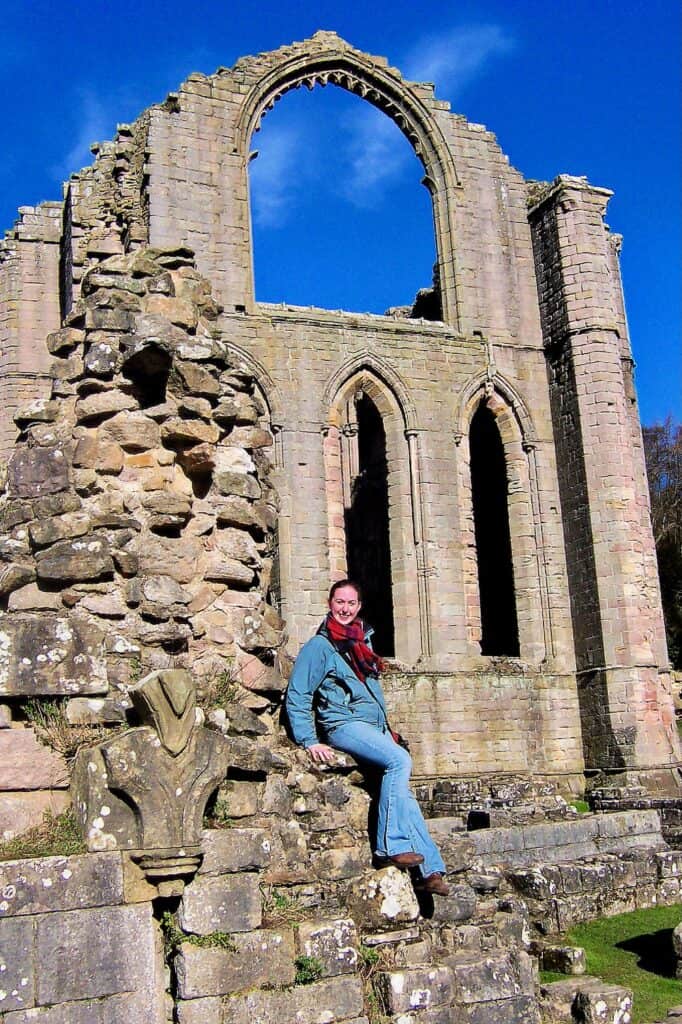 Woman perches on ruined stone wall in front of abbey ruins at Fountains Abbey Yorkshire