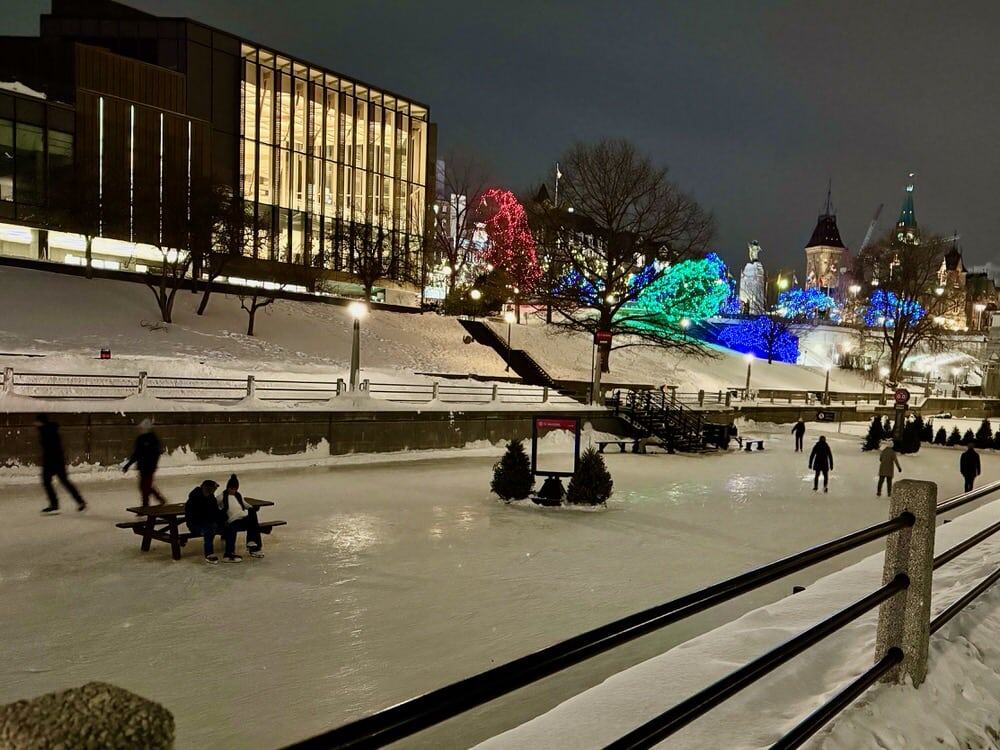 Skaters on the Rideau Canal after dark near the Confederation Park and the Houses of Parliament with Christmas lights in the background.