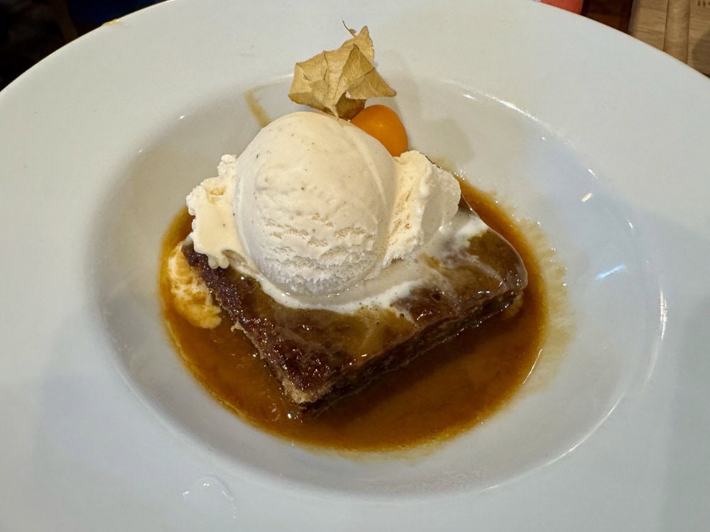 A white bowl filled with a piece of date cake covered with a toffee sauce and vanilla ice cream