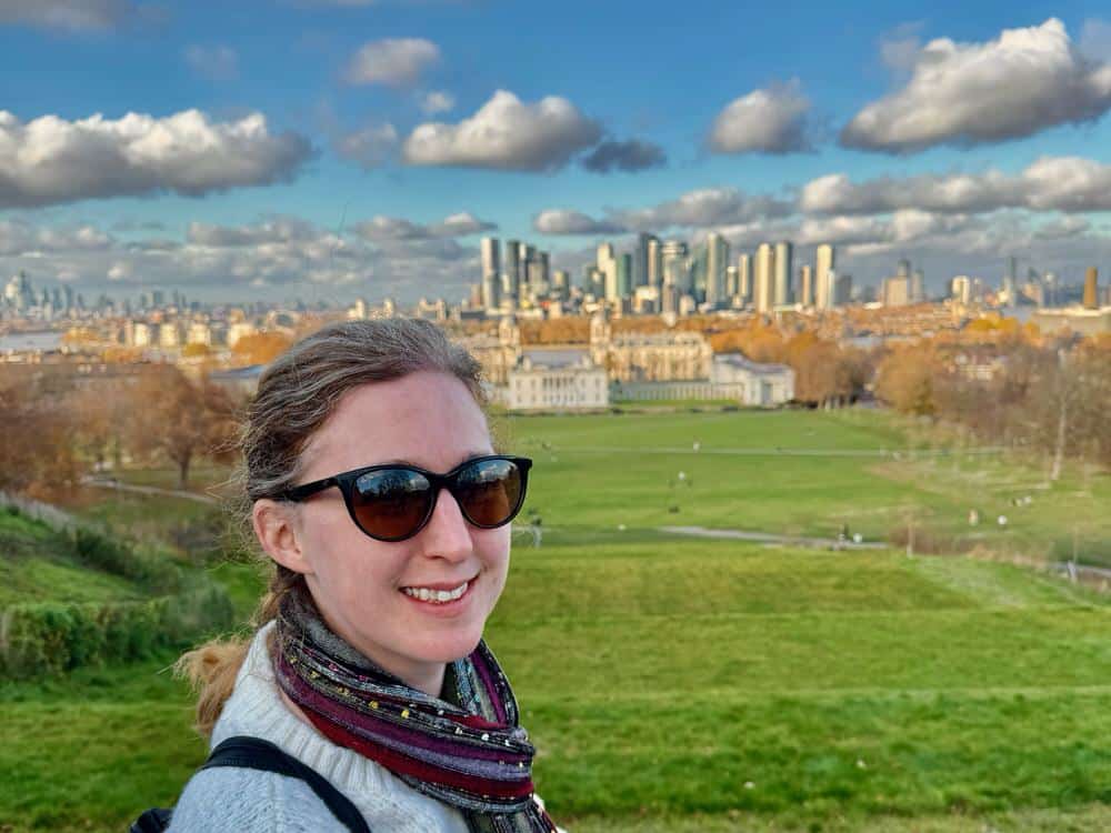 Woman poses at the top of Observatory Hill in front of a view of the Queen's House, Old Royal Naval College, and Canary Wharf in southeast London.