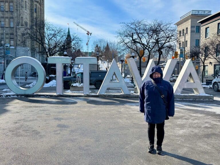 A woman bundled up in a winter parka poses in front of an Ottawa sign