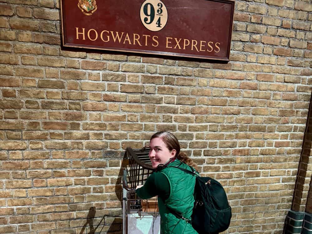 Woman poses with a trolley disappearing into a brick wall at the Platform 9 3/4 set at Warner Bros Studio London