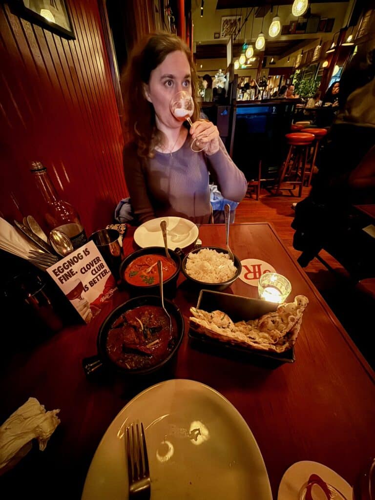 Woman sips her drink while sitting in a dark Indian restaurant in front of a table covered with Indian dishes