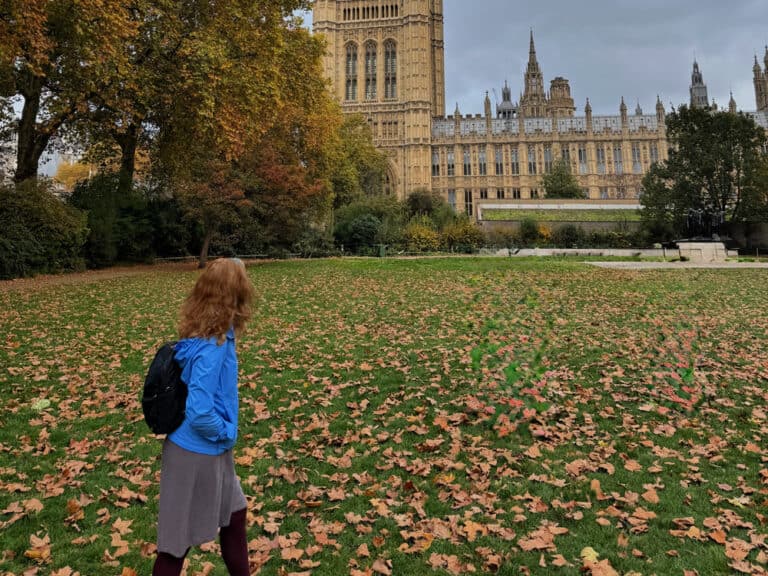 Woman wearing a rain jacket and backpack walks in autumn leaves behind the Houses of Parliament in London