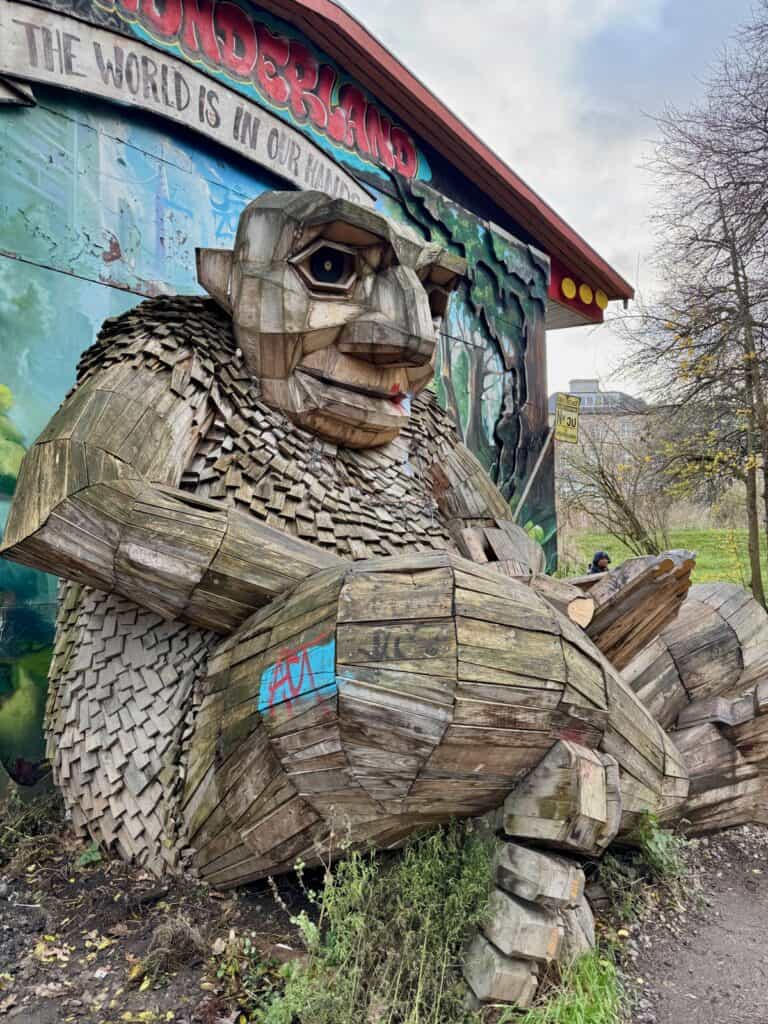 Large wooden troll statue sits in front of a mural on the side of a buildng