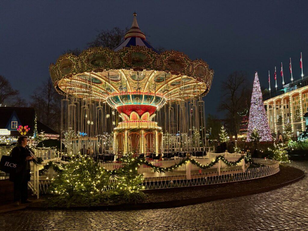 A woman stands next to a brightly liit carousel ride in Tivoli Gardens amusement park in Copenhagen Denmark