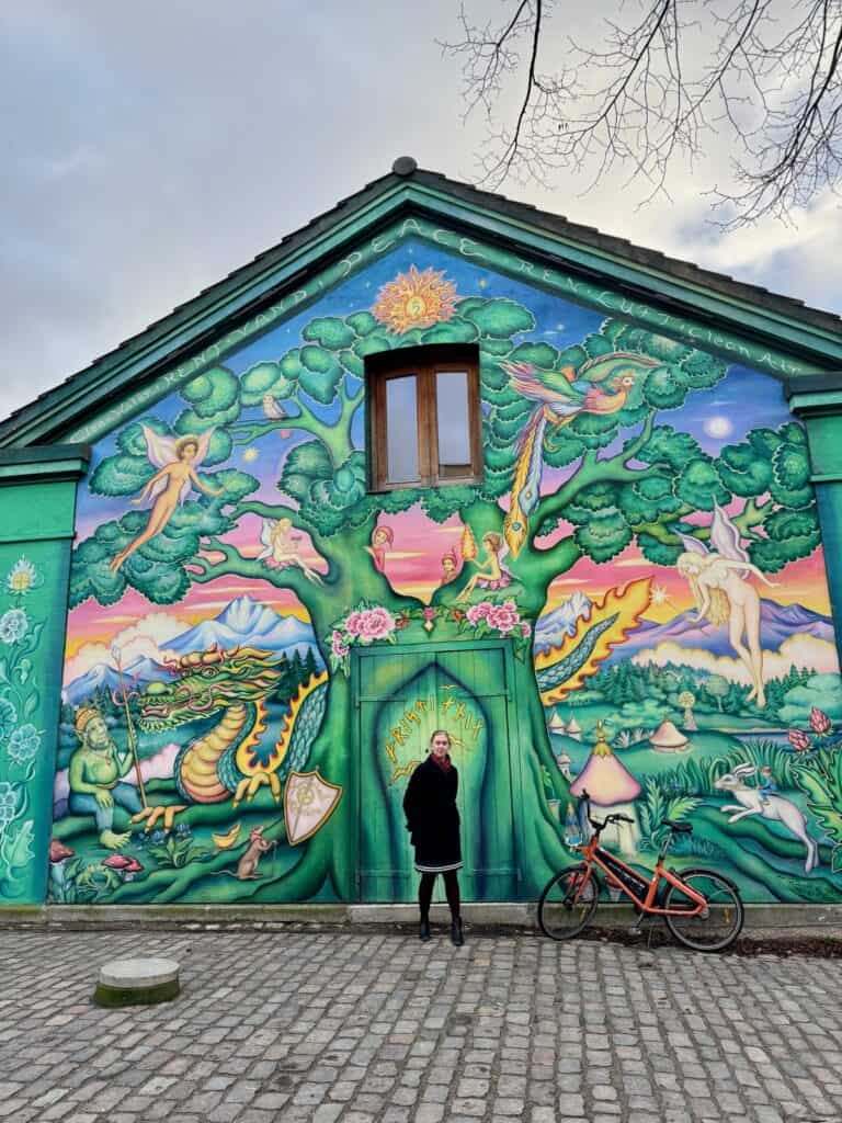 Woman poses in front of a mural depicting fairies and a fantasy scene on the side of a building.