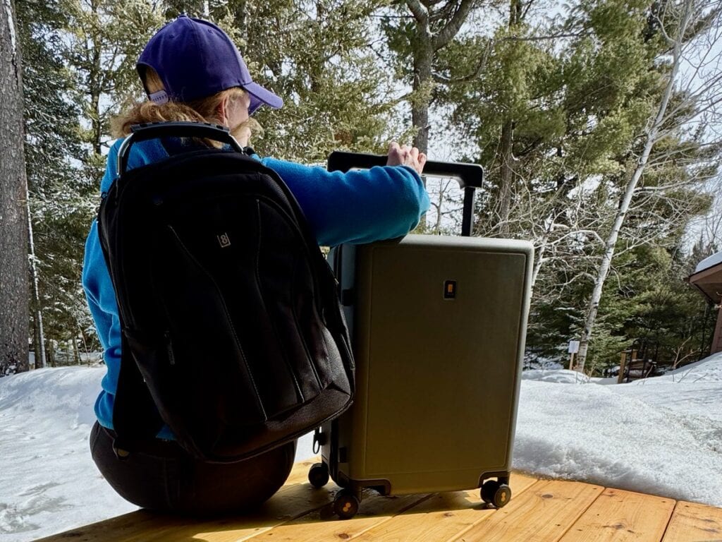 Woman sits with a LEVEL8 backpack on while holding the wide handle of a Voyageur suitcase