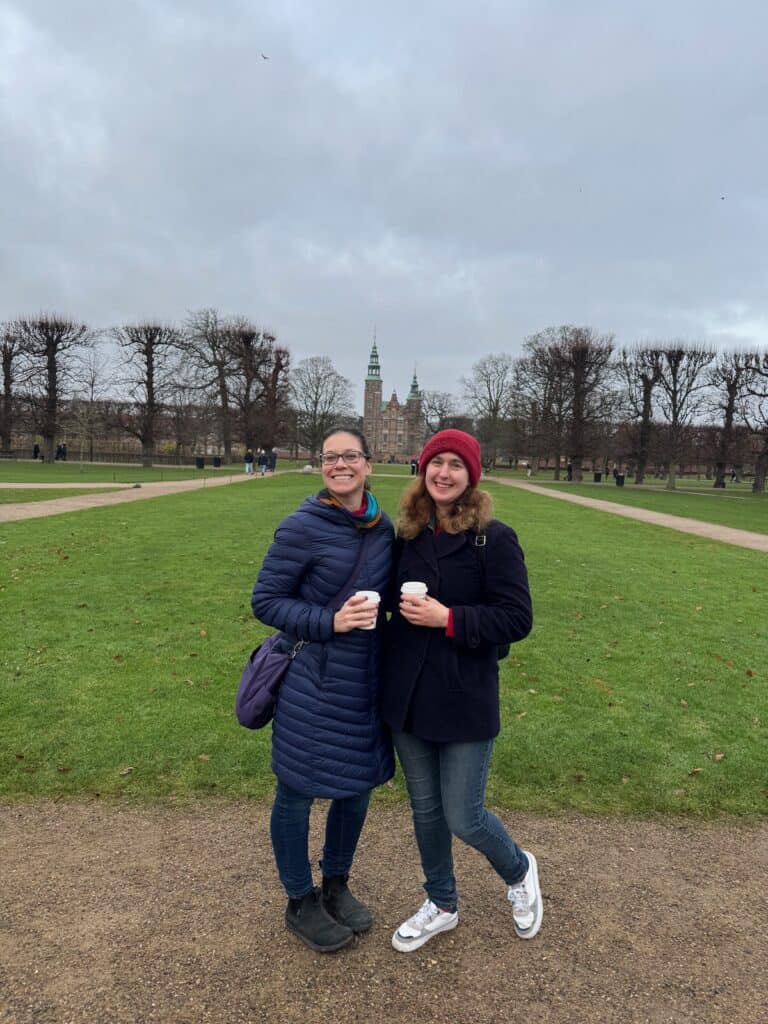 Two women holding coffee mugs pose in front of Rosenborg Slot in a city park