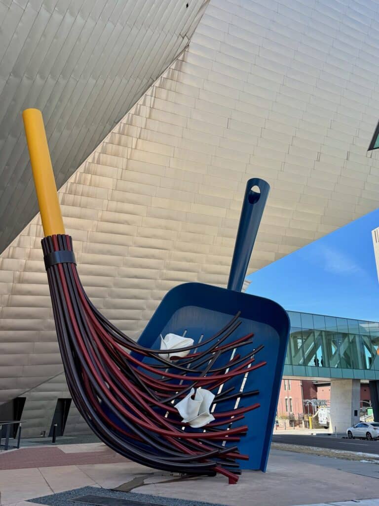 Sculpture of an oversized hand broom sweep up rubbish into a blue oversized dustpan outside the Denver Art Museum
