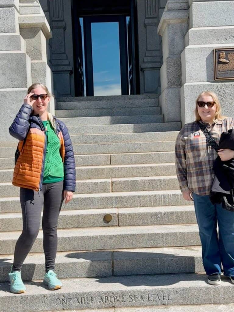 Two women stand on the steps of the Colorado State Capitol Building on the step engraved with "One Mile Above Sea Level")